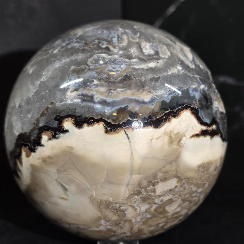 Peut inclure: Une sph&egrave;re polie &agrave; l'aspect marbr&eacute;. La pierre pr&eacute;sente un m&eacute;lange de couleurs grises, noires et cr&egrave;me, avec des motifs et des veines complexes. La sph&egrave;re est lisse et r&eacute;fl&eacute;chissante, mettant en valeur les variations naturelles.