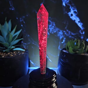 May include: A tall, red crystal point with a speckled pattern, mounted on a black, tiered base. The crystal is illuminated, showcasing its internal texture. Two potted succulents flank the crystal, set against a dark background.