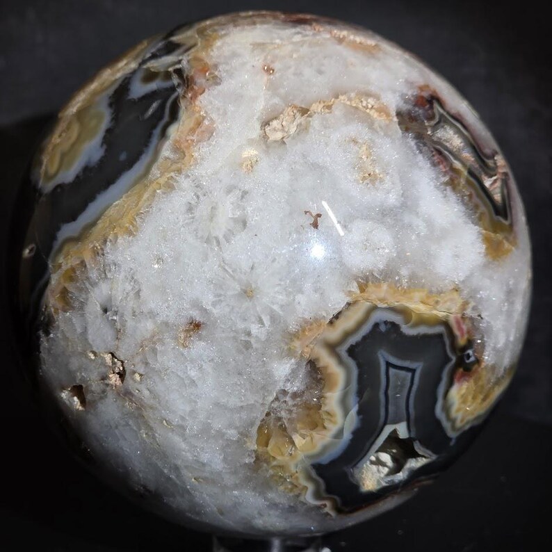 May include: A polished agate sphere with a mix of colors, including white, black, brown, and tan. The sphere has a natural, layered pattern and a smooth, reflective surface. This decorative sphere is a unique home decor item.