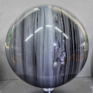 May include: A polished, spherical decorative object with a black and gray striped pattern. The sphere is displayed on a black stand, showcasing its reflective surface and unique design. The overall aesthetic is modern and elegant.
