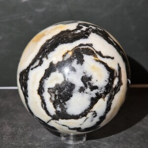 May include: A polished stone sphere featuring a high-contrast pattern of swirling black and white bands with subtle golden-yellow inclusions. The surface is smooth and reflective, showcasing organic, irregular mineral veins throughout the orb.