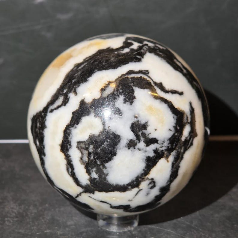 May include: A polished stone sphere featuring a high-contrast pattern of swirling black and white bands with subtle golden-yellow inclusions. The surface is smooth and reflective, showcasing organic, irregular mineral veins throughout the orb.