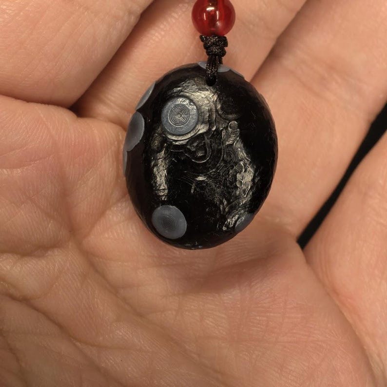 May include: A black, oval-shaped pendant with gray polka dots and a spiral design. The pendant is suspended from a black cord with a red bead at the top. The pendant is held in a person's hand.