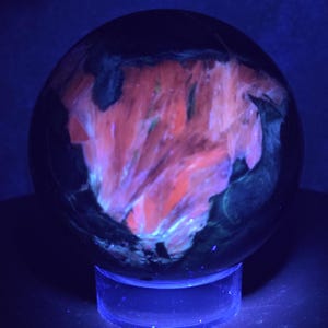 May include: A spherical mineral specimen resting on a clear circular stand. Under ultraviolet light, the stone displays a dark, opaque exterior with a prominent, glowing patch of vibrant orange and pink crystalline inclusions across the surface.