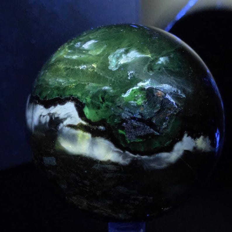 Peut inclure: Une pierre pr&eacute;cieuse sph&eacute;rique polie, pr&eacute;sentant un motif tourbillonnant de vert fonc&eacute;, de blanc et de noir. La surface est lisse et r&eacute;fl&eacute;chissante, avec des couleurs qui se fondent en un effet marbr&eacute;. L'objet a un aspect naturel, avec des tons terre.