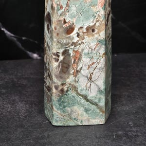 May include: A polished stone obelisk featuring a complex pattern of sage green, dark brown, and cream-colored mineral inclusions. The surface displays a natural, marbled texture with visible fractures and varied earthy tones throughout the structure.