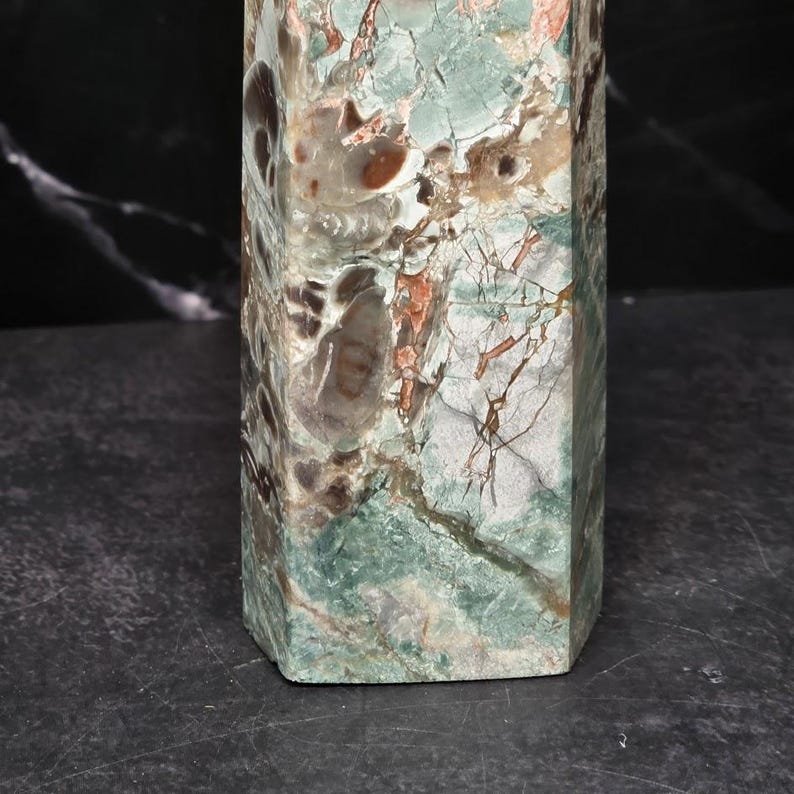 May include: A polished stone obelisk featuring a complex pattern of sage green, dark brown, and cream-colored mineral inclusions. The surface displays a natural, marbled texture with visible fractures and varied earthy tones throughout the structure.