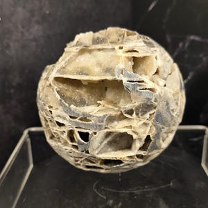 May include: A spherical, textured object with a lattice-like structure. The object is primarily beige and gray, with a network of openings throughout. The surface has a rough, uneven texture, and the overall shape is round.