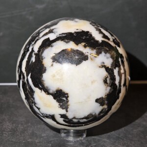 May include: A polished stone sphere featuring a high-contrast pattern of opaque white and deep black swirling veins. The surface is smooth and reflective, showcasing organic, irregular shapes that wrap around the entire spherical form.