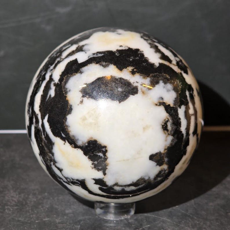 May include: A polished stone sphere featuring a high-contrast pattern of opaque white and deep black swirling veins. The surface is smooth and reflective, showcasing organic, irregular shapes that wrap around the entire spherical form.