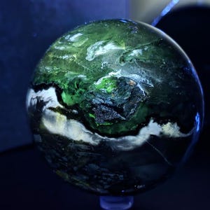May include: A spherical decorative object with a marbled appearance. The sphere features a blend of green, white, and black colors, creating a unique pattern. The object is displayed on a clear stand, and the background is dark.