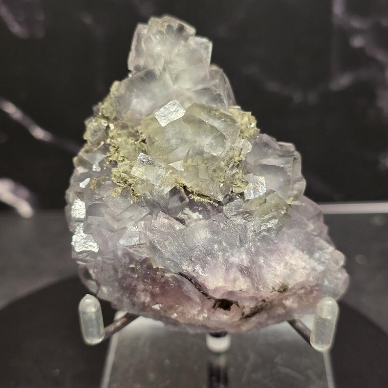 Puede incluir: Primer plano de un esp&eacute;cimen mineral de fluorita. La formaci&oacute;n cristalina presenta una mezcla de cristales c&uacute;bicos de color p&uacute;rpura p&aacute;lido y transparente con una capa de pirita dorada. El esp&eacute;cimen est&aacute; montado sobre un soporte transparente.