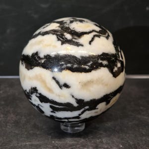 May include: A polished spherical stone featuring a high-contrast pattern of swirling black and creamy white bands. The surface is smooth and reflective, showcasing organic, flowing lines that wrap around the entire circumference of the sphere.