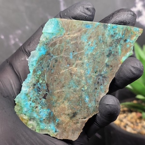 May include: A raw, polished stone slab with a mix of colors. The stone features a base of brown and gray with patches of vibrant turquoise and teal. The edges are rough and uneven, showcasing the natural form of the mineral.