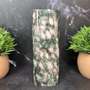 May include: A cylindrical stone sculpture featuring a mottled pattern of deep forest green and soft beige patches. The surface has a polished, smooth texture with visible mineral inclusions throughout the vertical, column-like structure.