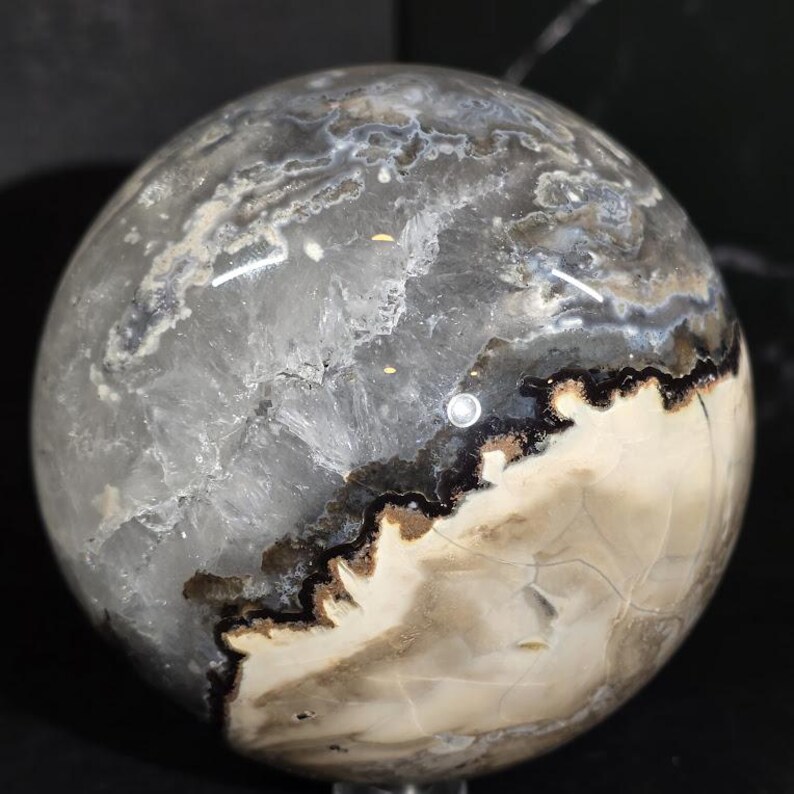 Peut inclure: Une sph&egrave;re polie &agrave; l'aspect marbr&eacute;. La pierre pr&eacute;sente un m&eacute;lange de tons gris, blancs et bruns, avec des veines brun fonc&eacute; et noires. La sph&egrave;re a une surface lisse et r&eacute;fl&eacute;chissante, probablement un objet d&eacute;coratif.