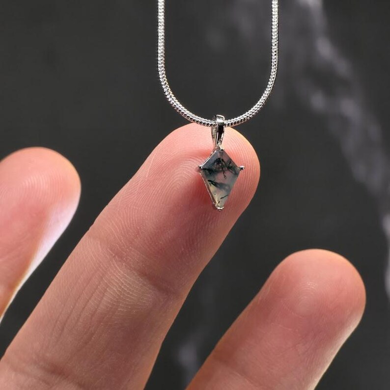 Puede incluir: Un collar plateado con un peque&ntilde;o colgante en forma de diamante. El colgante presenta un patr&oacute;n verde y blanco dentro de un marco plateado. El collar se sostiene entre dos dedos sobre un fondo oscuro.