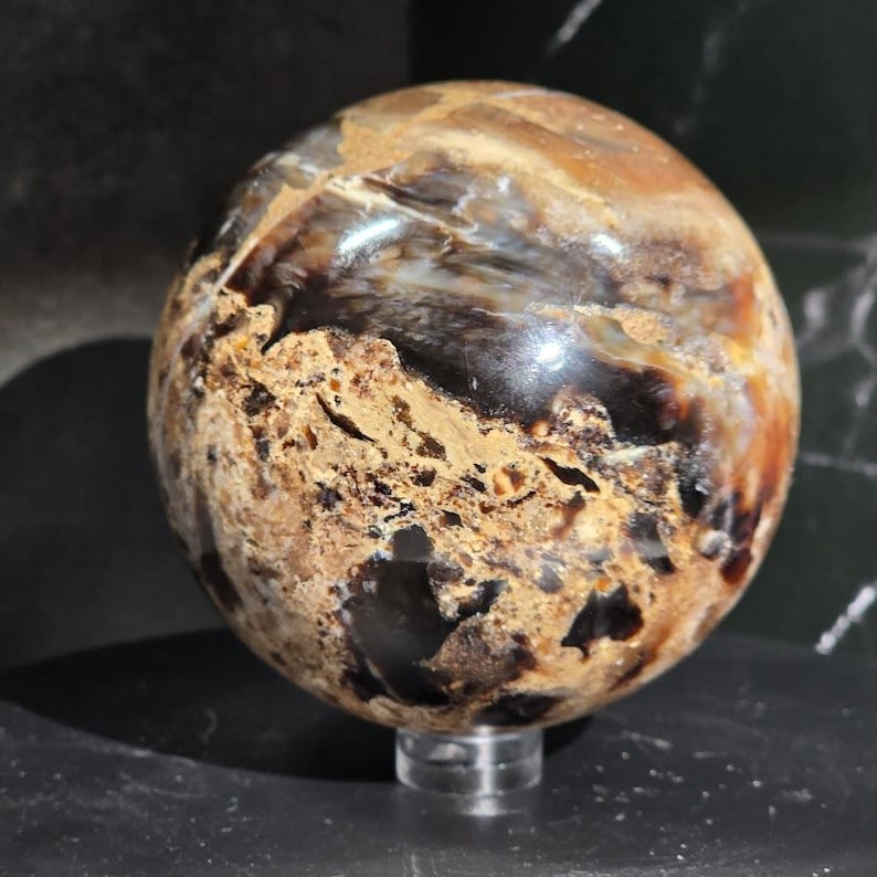 May include: A polished spherical stone featuring a complex pattern of deep brown, black, and tan earthy tones. The surface displays a mix of smooth, glossy areas and rough, textured patches with visible cracks and natural mineral inclusions.