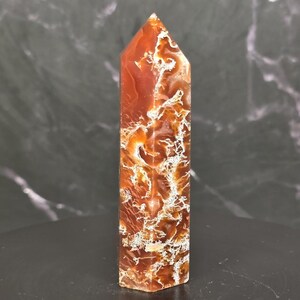 May include: A polished, six-sided crystal tower featuring a deep reddish-orange hue with intricate, branching white and translucent inclusions throughout the stone. The point is faceted into a sharp pyramid shape, showcasing a smooth, glossy finish.