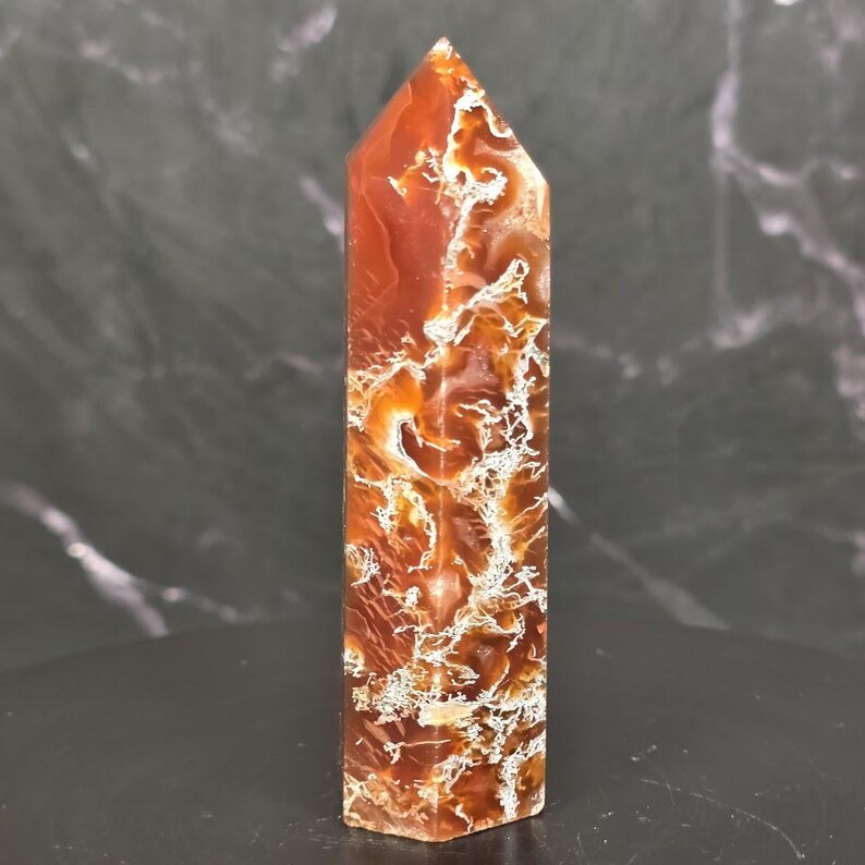 May include: A polished, six-sided crystal tower featuring a deep reddish-orange hue with intricate, branching white and translucent inclusions throughout the stone. The point is faceted into a sharp pyramid shape, showcasing a smooth, glossy finish.