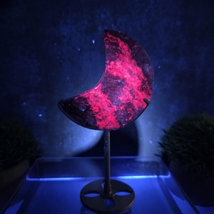 May include: A crescent moon-shaped mineral specimen featuring a dark, mottled surface with vibrant, glowing neon pink patches. The stone is held upright by a thin, dark metal stand with a circular base, illuminated under ultraviolet light.