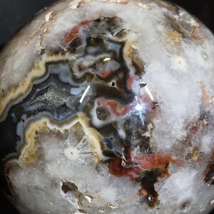 May include: A polished agate sphere with a complex pattern of colors. The sphere features bands of white, black, brown, red, and tan hues. The surface has a glossy finish, and the overall shape is spherical.