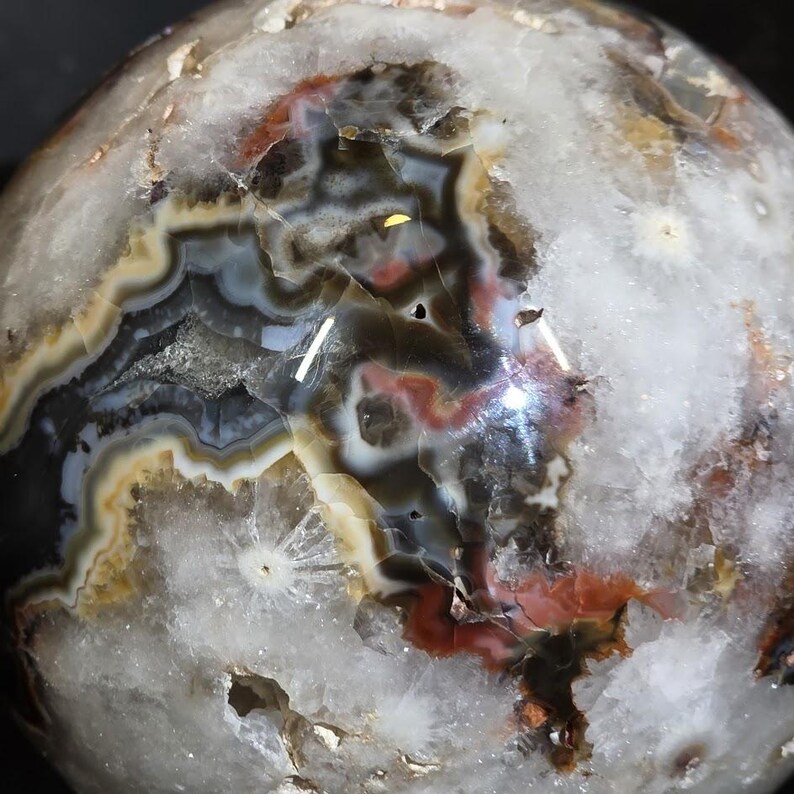May include: A polished agate sphere with a complex pattern of colors. The sphere features bands of white, black, brown, red, and tan hues. The surface has a glossy finish, and the overall shape is spherical.