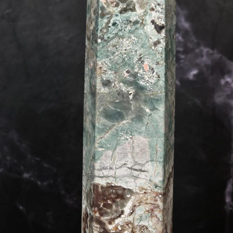 May include: A tall, hexagonal crystal tower featuring a polished surface with a marbled appearance. The stone displays a blend of pale mint green, deep forest green, and earthy brown tones, characterized by intricate white veining and fissures.