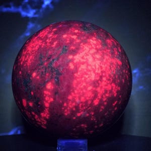 May include: A spherical stone displaying a vibrant, glowing red fluorescence across its surface, contrasted by dark, irregular mineral inclusions. The object exhibits a mottled texture with bright crimson patches that appear to emit light.