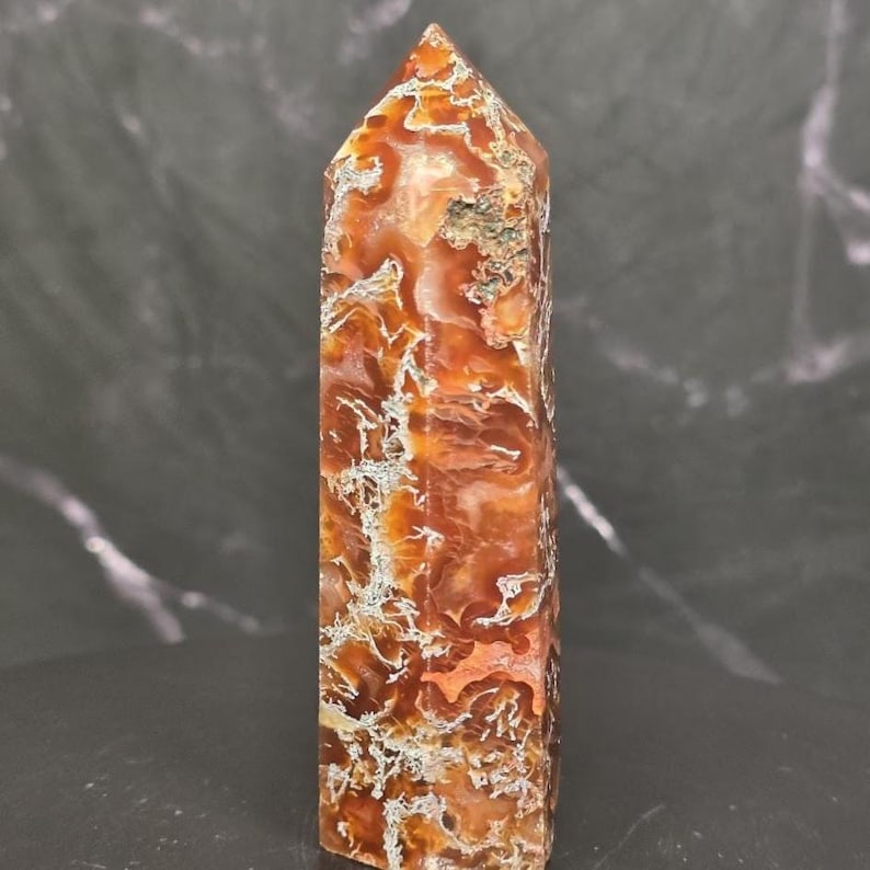 May include: A polished stone tower with a hexagonal point, featuring a vibrant mix of burnt orange, deep amber, and translucent brown hues. The surface displays intricate, web-like white and grey mineral inclusions throughout the crystalline structure.