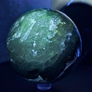 Peut inclure: Une pierre pr&eacute;cieuse sph&eacute;rique translucide avec des motifs cristallins verts et blancs. La surface polie refl&egrave;te la lumi&egrave;re, cr&eacute;ant un effet lumineux. La sph&egrave;re est plac&eacute;e sur un fond sombre, soulignant sa beaut&eacute; naturelle.