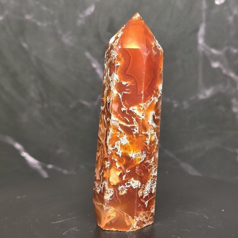 May include: A polished, hexagonal crystal tower featuring a vibrant, translucent orange and deep red color palette. The stone displays intricate, web-like white inclusions and natural fractures throughout its structure, ending in a sharp point.