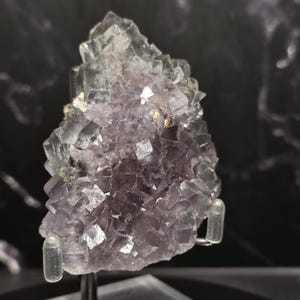 Puede incluir: Un grupo de cristales de fluorita natural, que muestra una gradaci&oacute;n de tonos morados. La estructura cristalina es visible, con algunas &aacute;reas transl&uacute;cidas. La muestra est&aacute; montada sobre un soporte transparente.