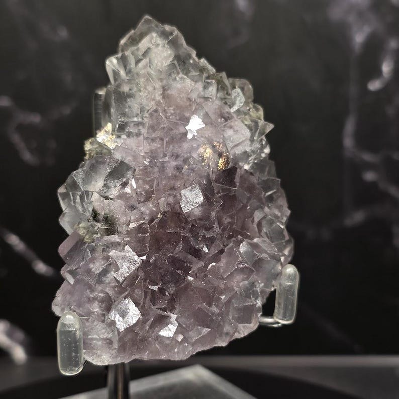 Puede incluir: Un grupo de cristales de fluorita natural, que muestra una gradaci&oacute;n de tonos morados. La estructura cristalina es visible, con algunas &aacute;reas transl&uacute;cidas. La muestra est&aacute; montada sobre un soporte transparente.