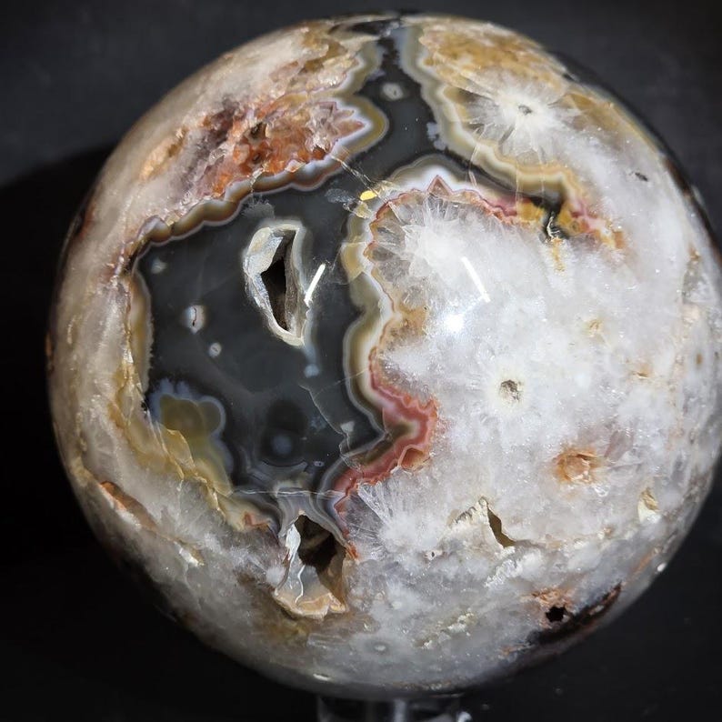 May include: A polished agate sphere with a geode-like interior. The sphere features a mix of colors, including white, gray, brown, and red. The surface has a polished, smooth texture, and the interior reveals crystalline formations.