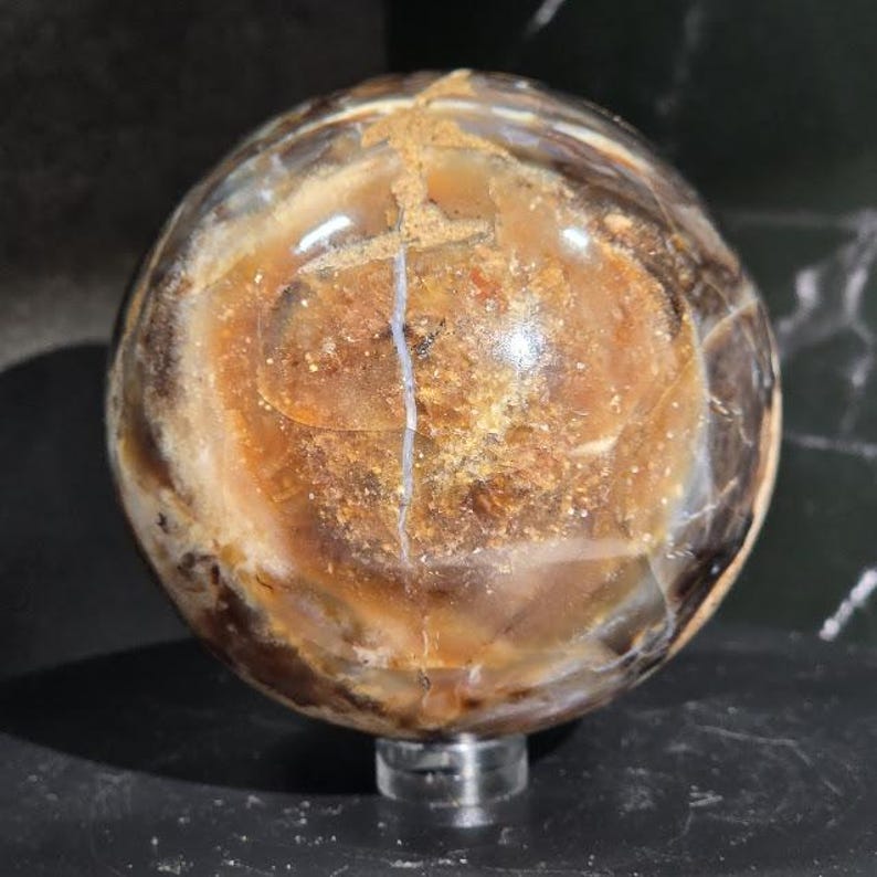 May include: A polished, spherical gemstone with a mix of brown, tan, and white hues. The orb has a crack running down the center and is displayed on a clear, round stand. The stone's surface has a glossy finish.
