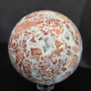 May include: A polished sphere of agate stone, featuring a mix of white, peach, and reddish-brown hues. The stone has a speckled pattern and a geode-like cavity. The sphere is displayed on a clear stand, showcasing its natural beauty.