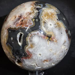 May include: A polished, spherical agate geode. The orb features a complex pattern of gray, white, brown, and gold bands and crystalline formations. The geode has several natural cavities and a glossy finish, showcasing its unique geological composition.
