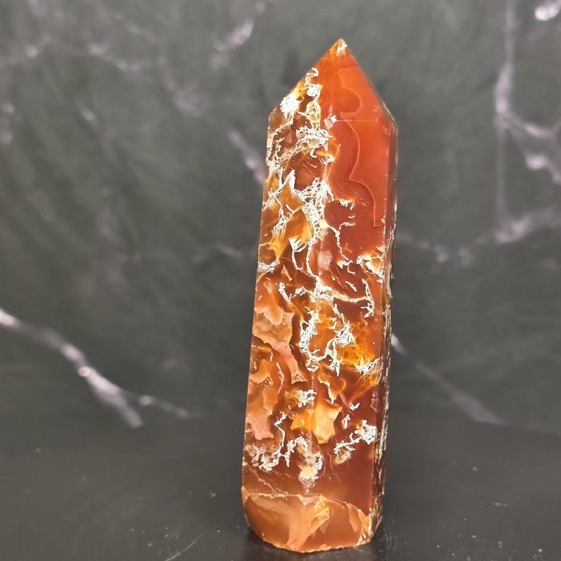 May include: A polished carnelian crystal tower with a hexagonal point. The stone features a deep burnt orange and reddish-brown color palette with intricate, web-like white inclusions and translucent patches throughout the vertical structure.