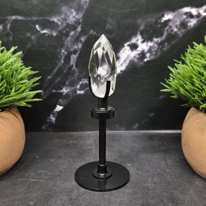 May include: A clear, faceted, pointed crystal stone is securely held in a black metal pronged mount. The crystal is supported by a slender black metal rod attached to a circular black base, creating a vertical display stand for the stone.