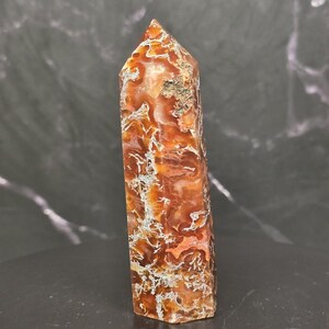 May include: A polished, six-sided crystal tower featuring a vibrant mix of burnt orange, deep red, and translucent amber hues. The stone displays intricate, web-like white and grey mineral inclusions throughout its vertical, faceted structure.