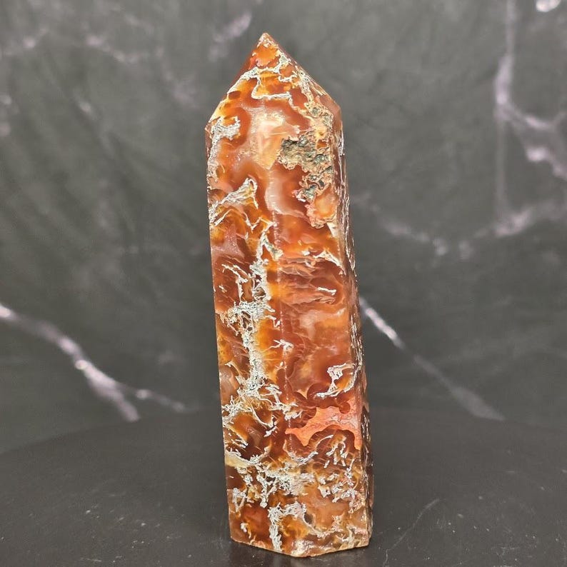 May include: A polished, six-sided crystal tower featuring a vibrant mix of burnt orange, deep red, and translucent amber hues. The stone displays intricate, web-like white and grey mineral inclusions throughout its vertical, faceted structure.
