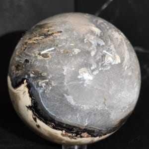 Peut inclure: Une boule de cristal sph&eacute;rique transparente avec des motifs internes blancs, noirs et bruns. La surface polie refl&egrave;te la lumi&egrave;re, mettant en valeur les formations min&eacute;rales naturelles. La sph&egrave;re est un objet d&eacute;coratif.