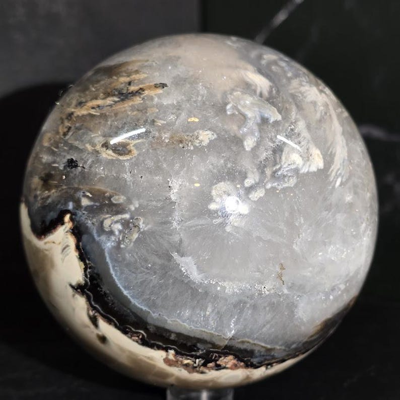 Peut inclure: Une boule de cristal sph&eacute;rique transparente avec des motifs internes blancs, noirs et bruns. La surface polie refl&egrave;te la lumi&egrave;re, mettant en valeur les formations min&eacute;rales naturelles. La sph&egrave;re est un objet d&eacute;coratif.