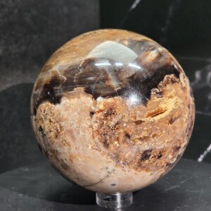 May include: A polished sphere with a mix of brown, tan, and black hues. The orb has a smooth, reflective surface with visible patterns and textures. It sits on a clear, small stand, showcasing its spherical shape. The overall aesthetic suggests a decorative or collectible item.