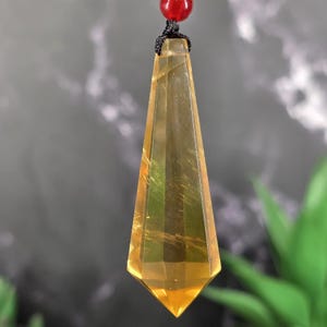 May include: A faceted, elongated pendant in shades of yellow and gold, suspended from a black cord and a red bead. The crystal pendant has a pointed bottom and a geometric design, with visible internal striations.