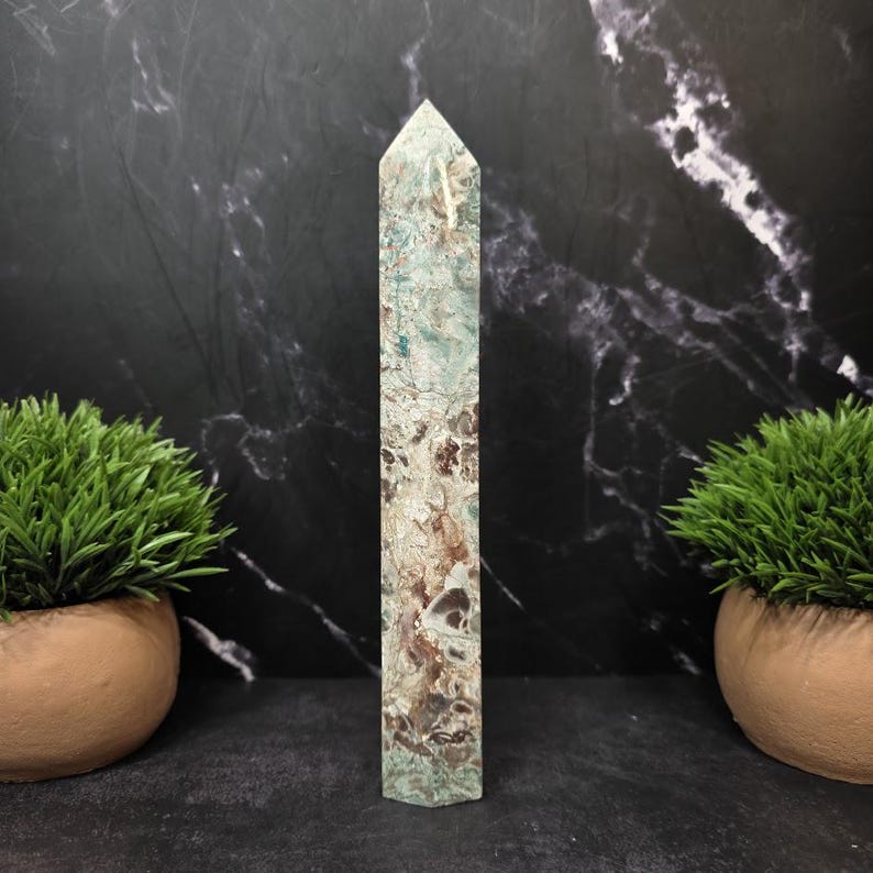 May include: A tall, six-sided crystal tower featuring a mottled pattern of sage green, cream, and dark brown mineral inclusions. The stone has a smooth, polished finish and terminates in a sharp, faceted point at the top, standing vertically.