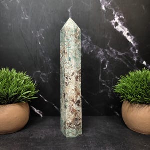 May include: A tall, six-sided crystal tower with a pointed top. The stone features a mottled pattern of light mint green, earthy brown, and creamy white colors throughout its surface, displaying a natural, polished, and smooth texture.