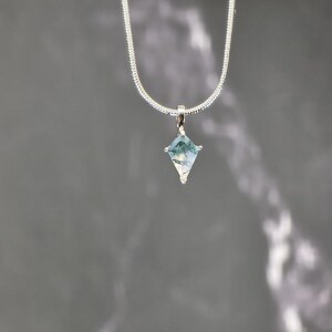 Puede incluir: Un collar de plata con un colgante en forma de diamante. El colgante presenta una piedra transparente con inclusiones verdes y blancas. La cadena es una fina cadena de serpiente plateada.