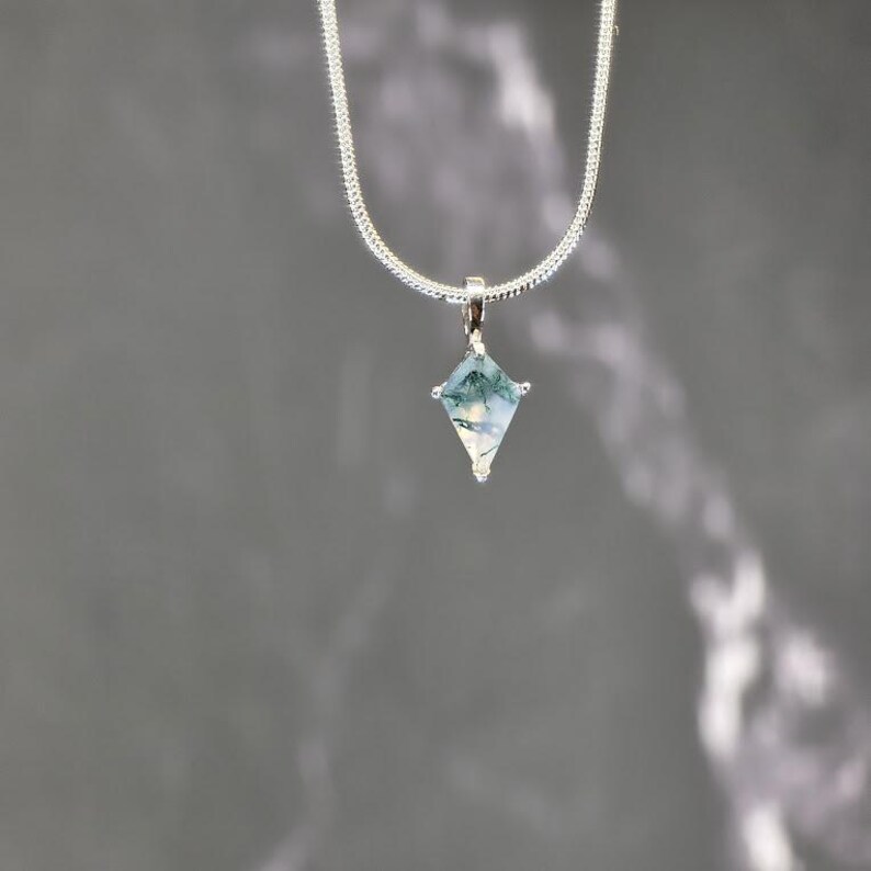 Puede incluir: Un collar de plata con un colgante en forma de diamante. El colgante presenta una piedra transparente con inclusiones verdes y blancas. La cadena es una fina cadena de serpiente plateada.